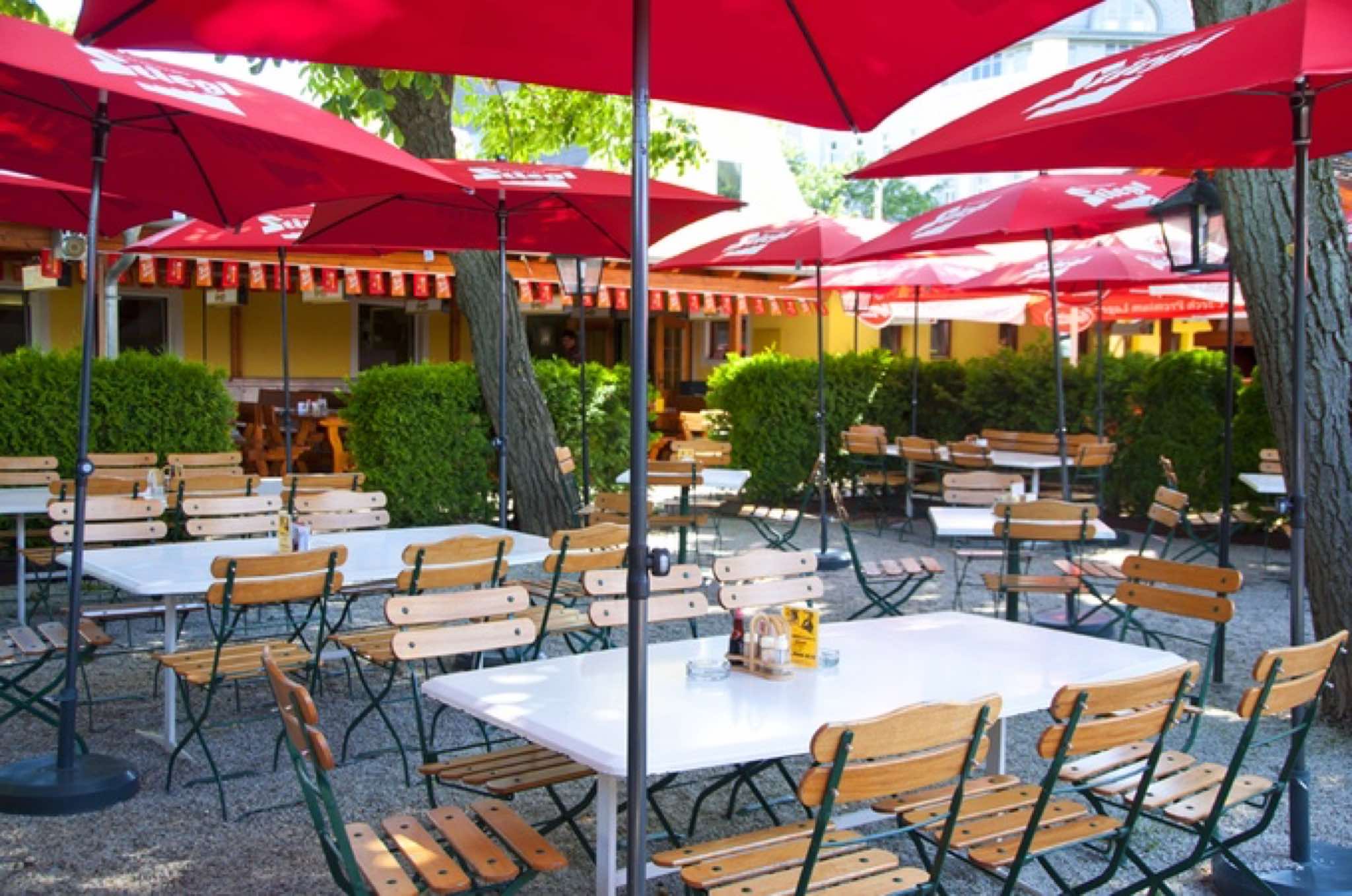 Gastgarten des Brigittenauer Stadl mit roten Sonnenschirmen und Bäumen